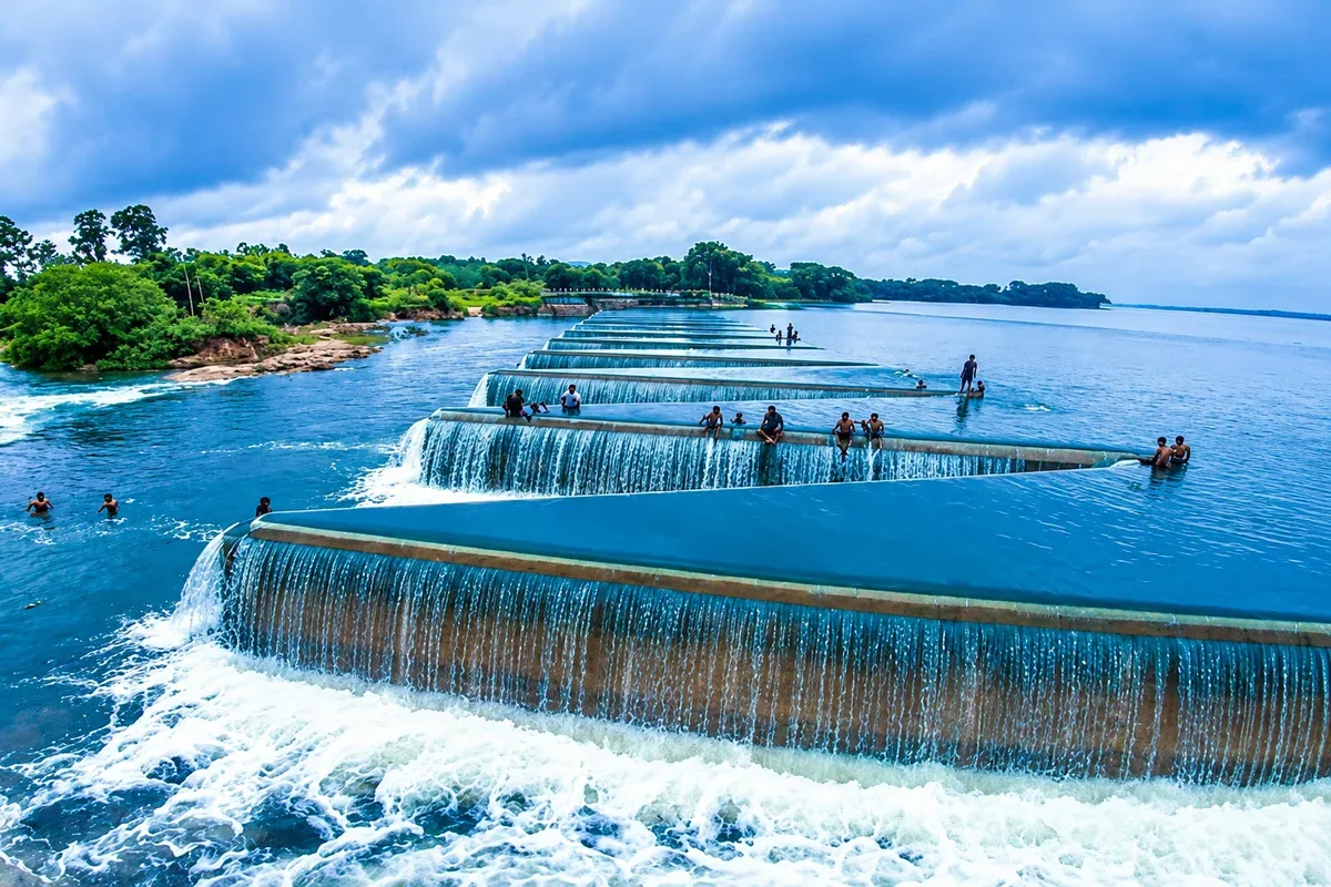 Water flowing over a long reservoir spillway with trees along the shore